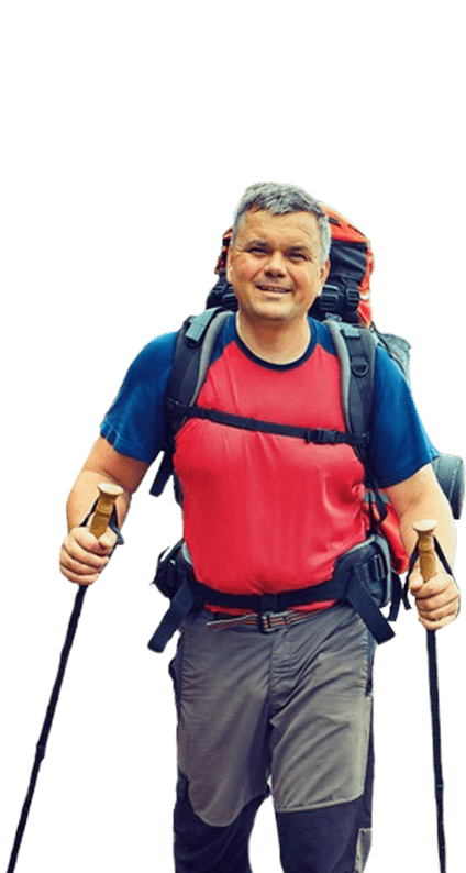 A man with a backpack and poles stands on a trail, surrounded by trees and mountains.